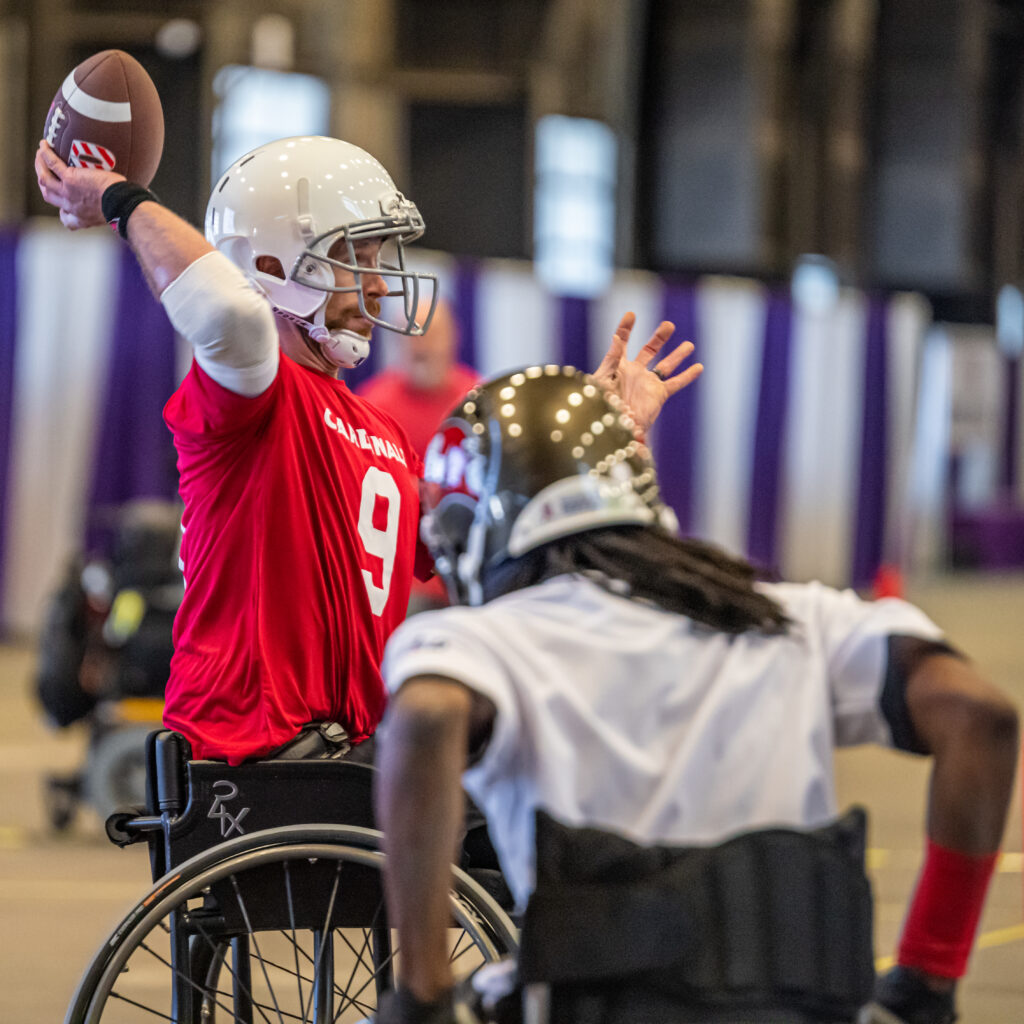 An Arizona Cardinal wheelchair football player is about to unleash a pass as a player on the opposing team charges.