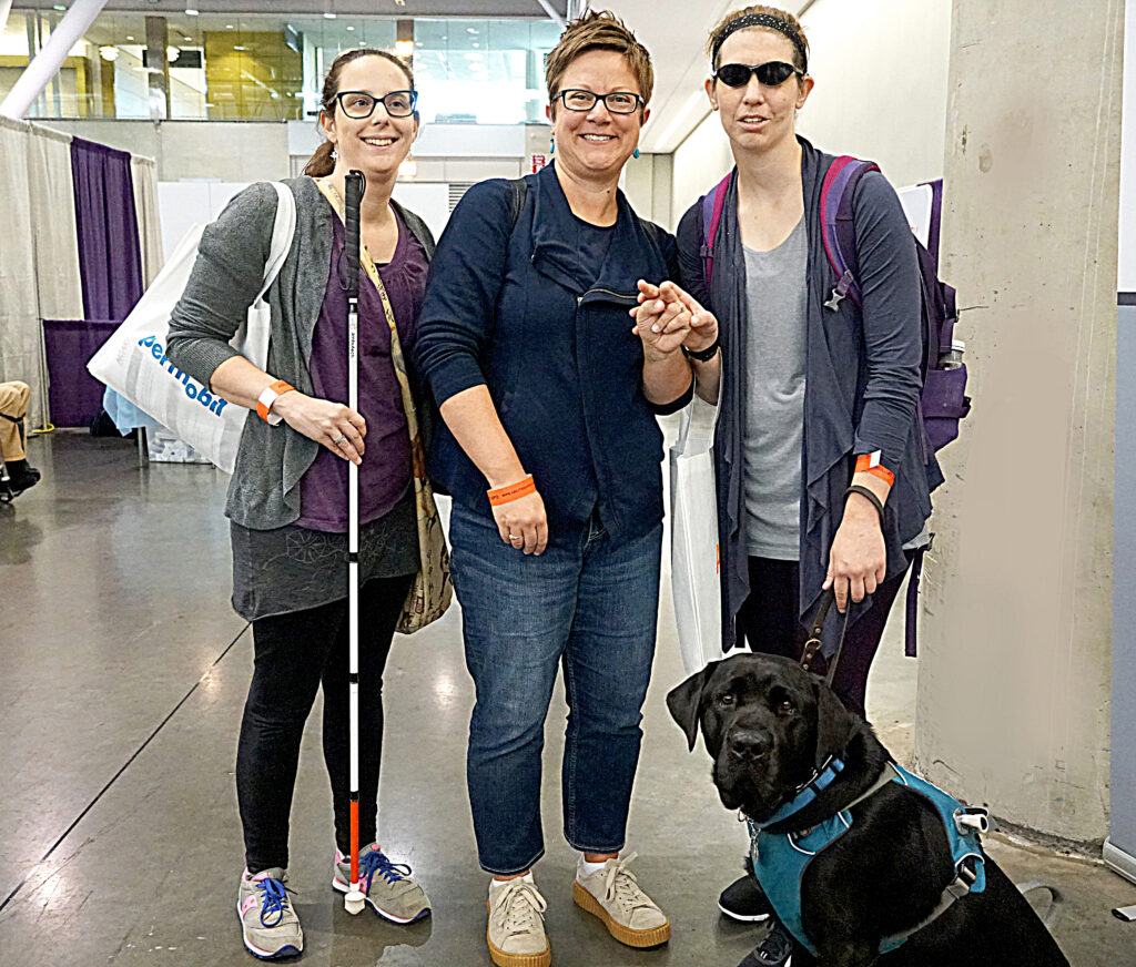 Of the three smiling Abilities Expo attendees, two are blind/low vision. The woman on the left is holding a white cane and the man on the rignt is wearing sun glasses and has a black service dog. 