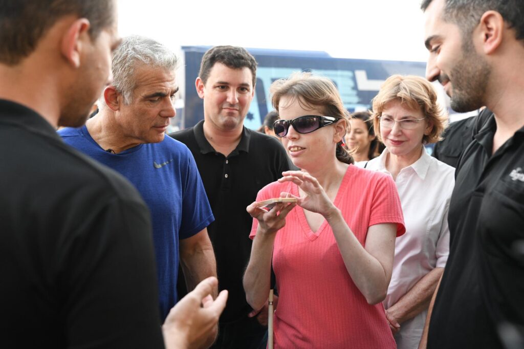 Photo of a woman who is either blind or low vision, talking to someone in a group. She is wearing a pink tshirt, and wearing black glasses