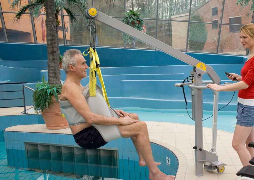 Man seated in a SureHands sling-style pool lift while an attendant operates the controls beside an indoor pool.