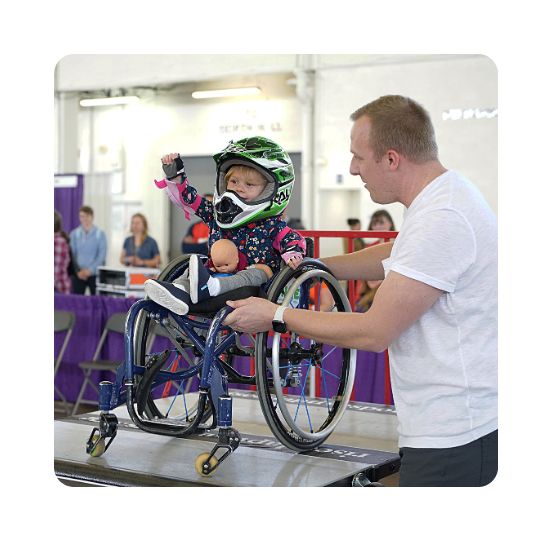 A little girl in a helmet raises her fist in triumph as she is about to go down a ramp in her wheelchair. Her dad is standing beside her and steadying the chair. 