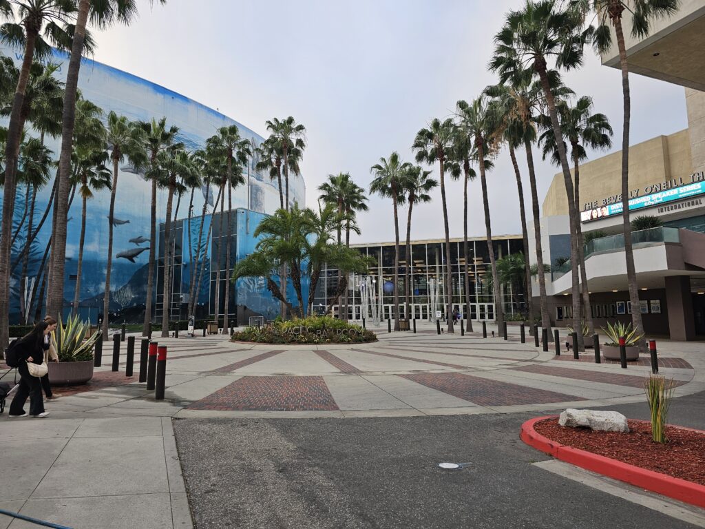 Wide view of the Long Beach Convention & Entertainment Center entrance, framed by tall palm trees. A large blue ocean-themed mural with whales and sea life covers the building on the left, while glass doors and signage for the Pacific Ballroom are centered. A few people walk along the paved plaza with brick accents and landscaped planters under an overcast sky.