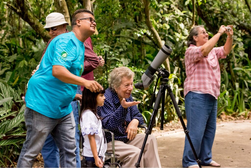 Group of people of different ages and abilities exploring a lush garden, with an older woman in a wheelchair smiling beside a spotting scope while others look and point at something in the trees.