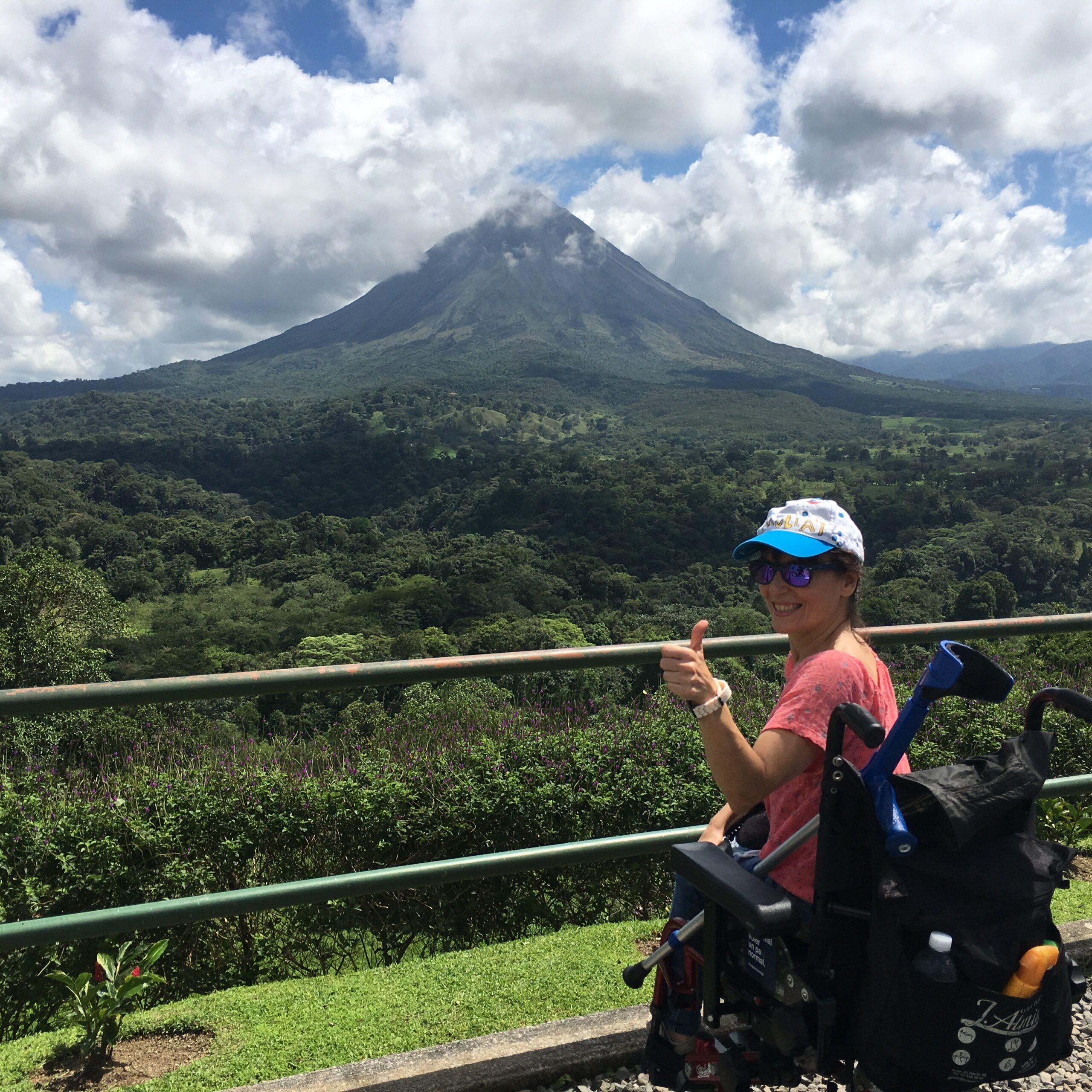 Woman using a wheelchair smiles and gives a thumbs up while overlooking a lush landscape with a volcano rising in the background.