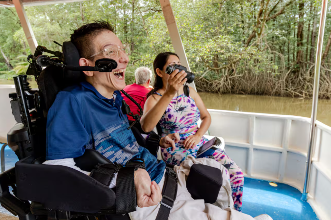 Man in a power wheelchair smiling and laughing on a small boat tour, while a woman next to him looks through binoculars, surrounded by a wooded waterway.