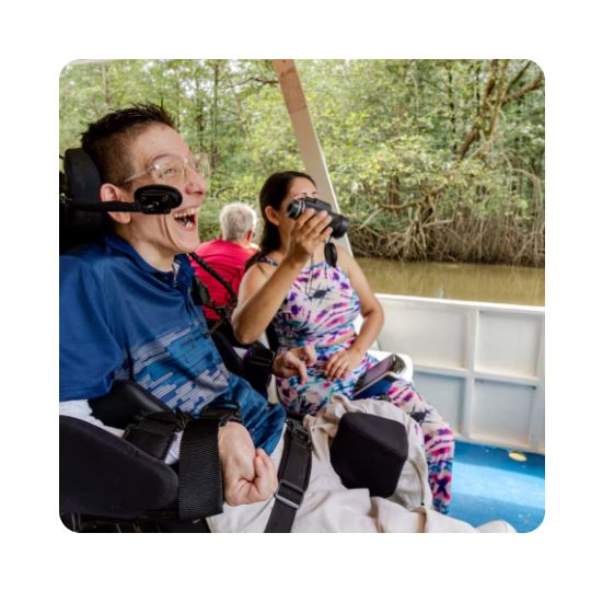 Man in a power wheelchair smiling and laughing on a small boat tour, while a woman next to him looks through binoculars, surrounded by a wooded waterway.