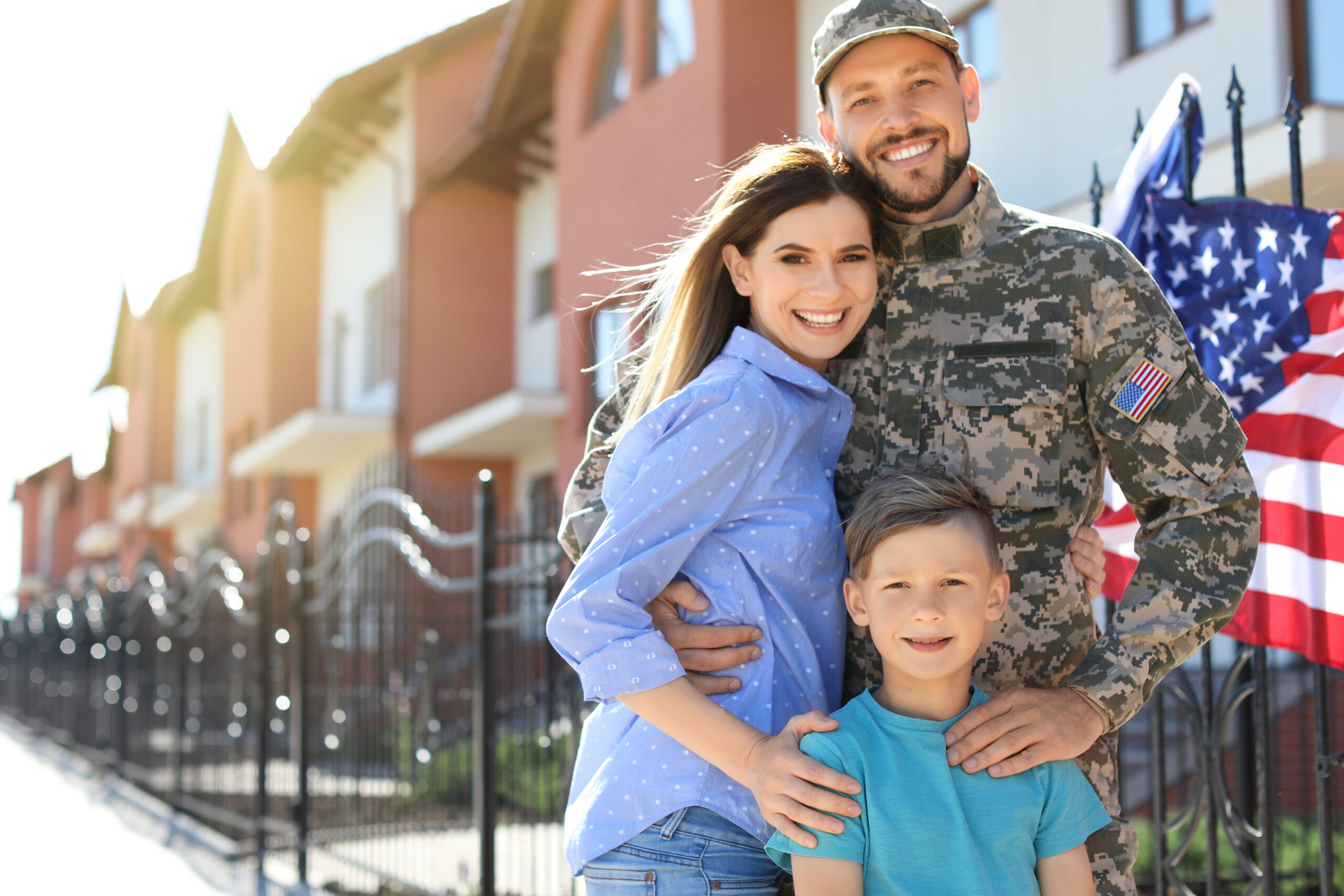Smiling military service member in uniform standing outside with partner and young child, an American flag visible in the background.
