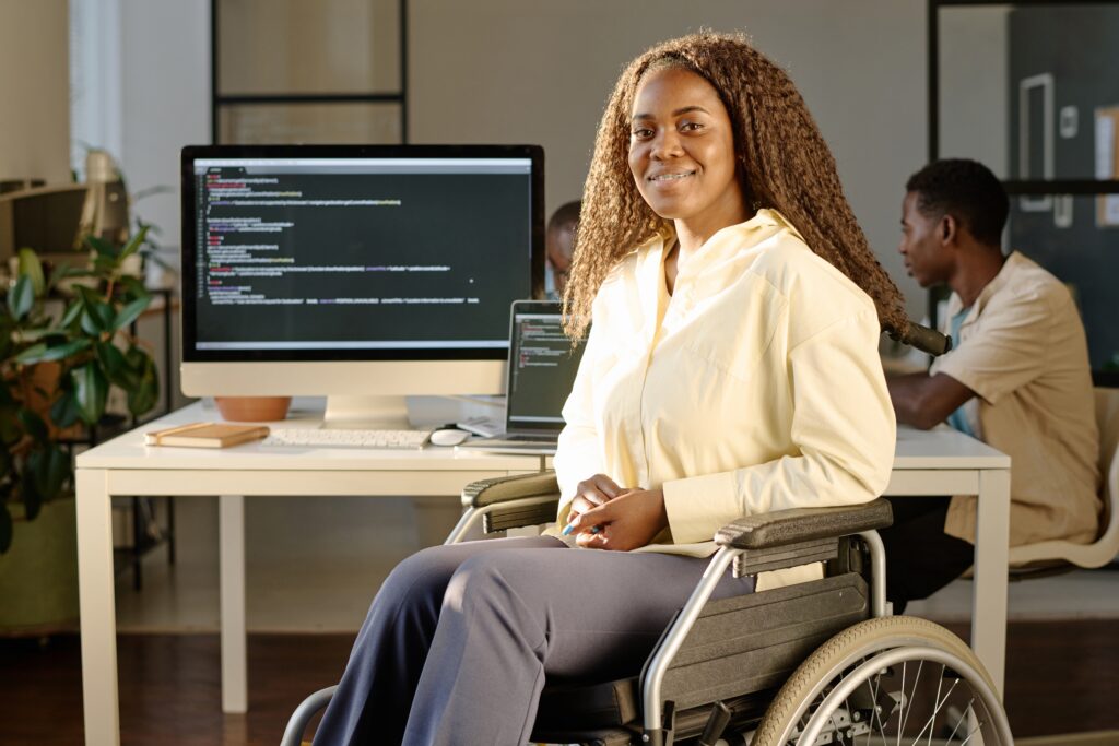 Young woman using a wheelchair smiling at the camera in a modern office, with computer screens showing code and coworkers in the background.