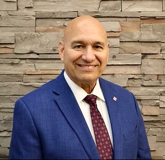 Headshot of John Becker with AMDA. He is smiling at the camera, wearing a blue suit jacket, white button up and maroon tie.