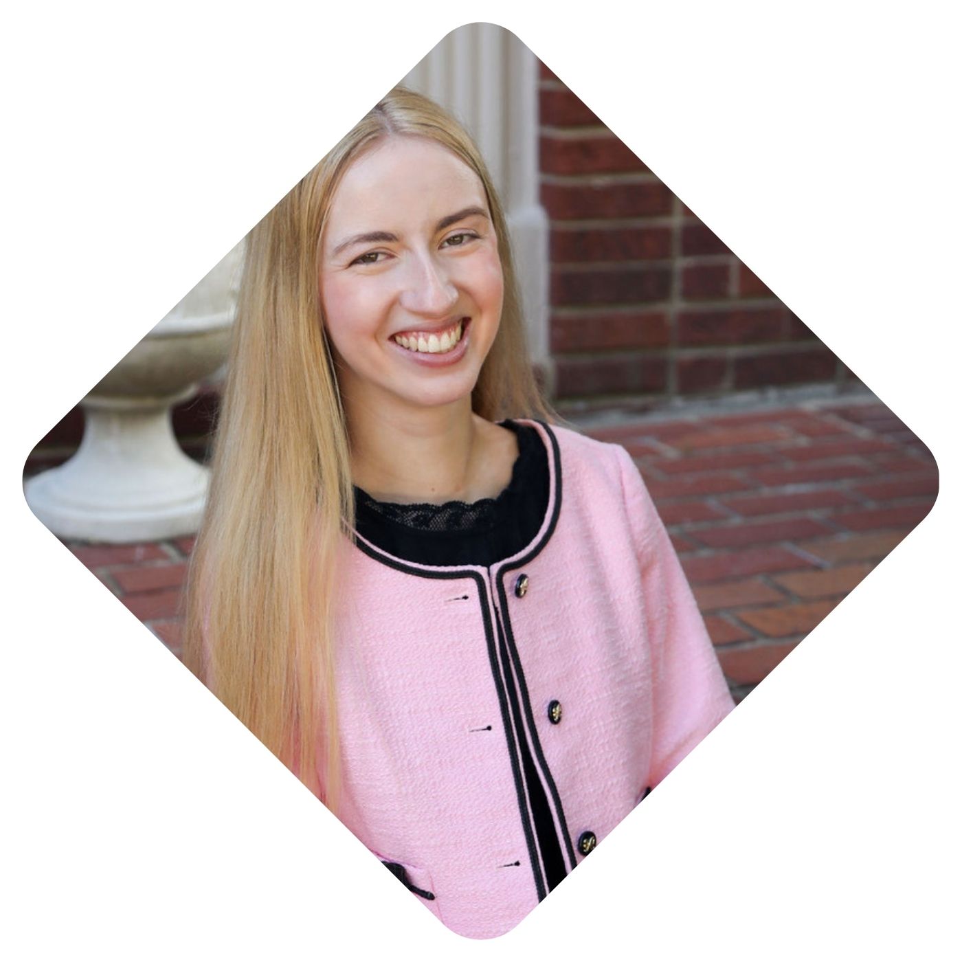 Headshot of Sarah Todd Hammer. She is sitting outside, wearing a black dress with a baby pink jacket. Her long blonde hair is worn down, and she is smiling at the camera.