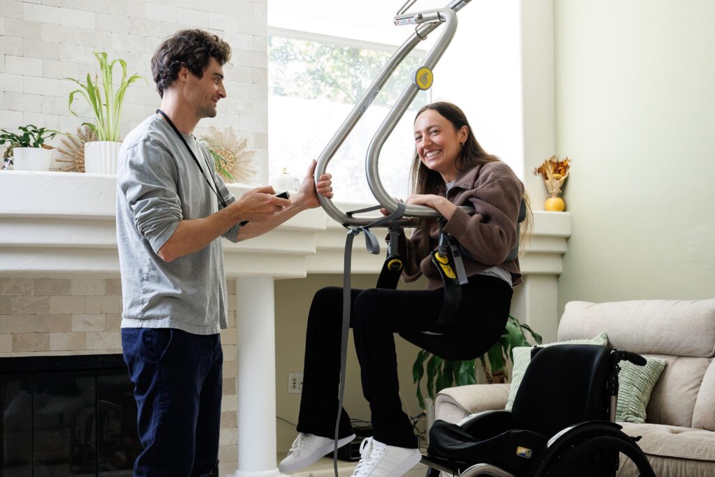 Woman using a SureHands lift with assistance from a caregiver in a bright living room, transferring safely from a wheelchair.