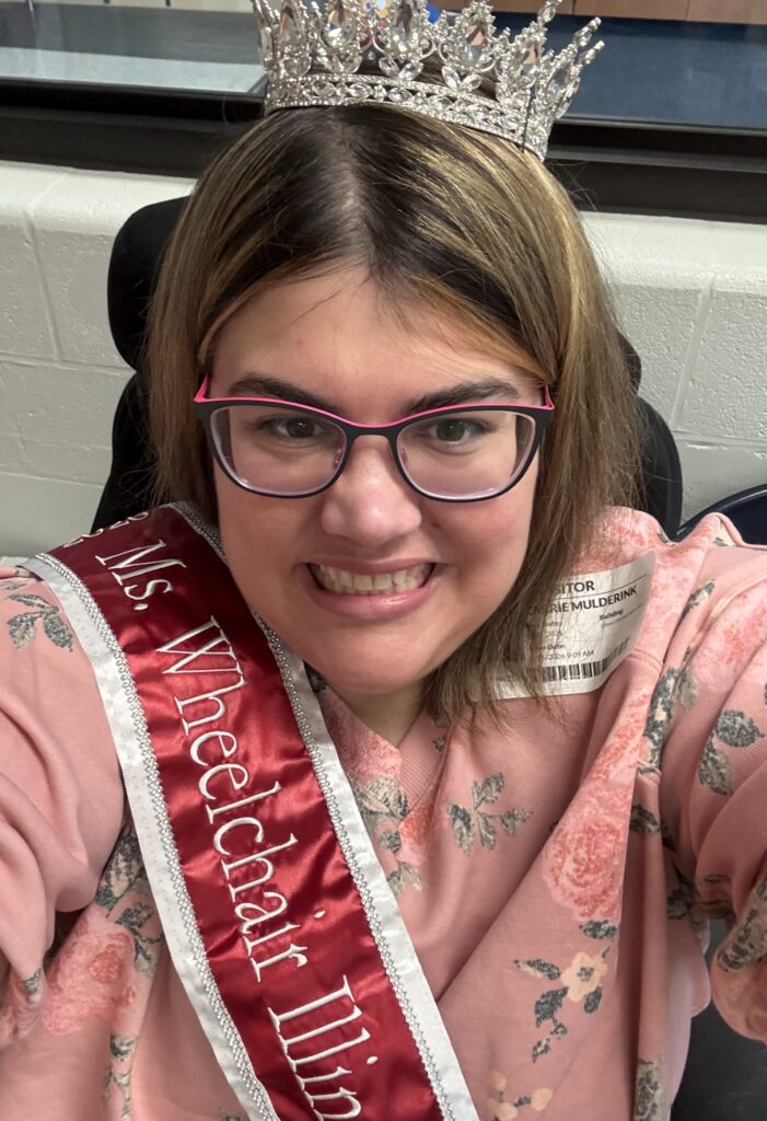 Ms. Wheelchair Illinois, Carrie Mulderink, takes a selfie in a pink dress, a tiara, and a Ms. Wheelchair Illinois sash.