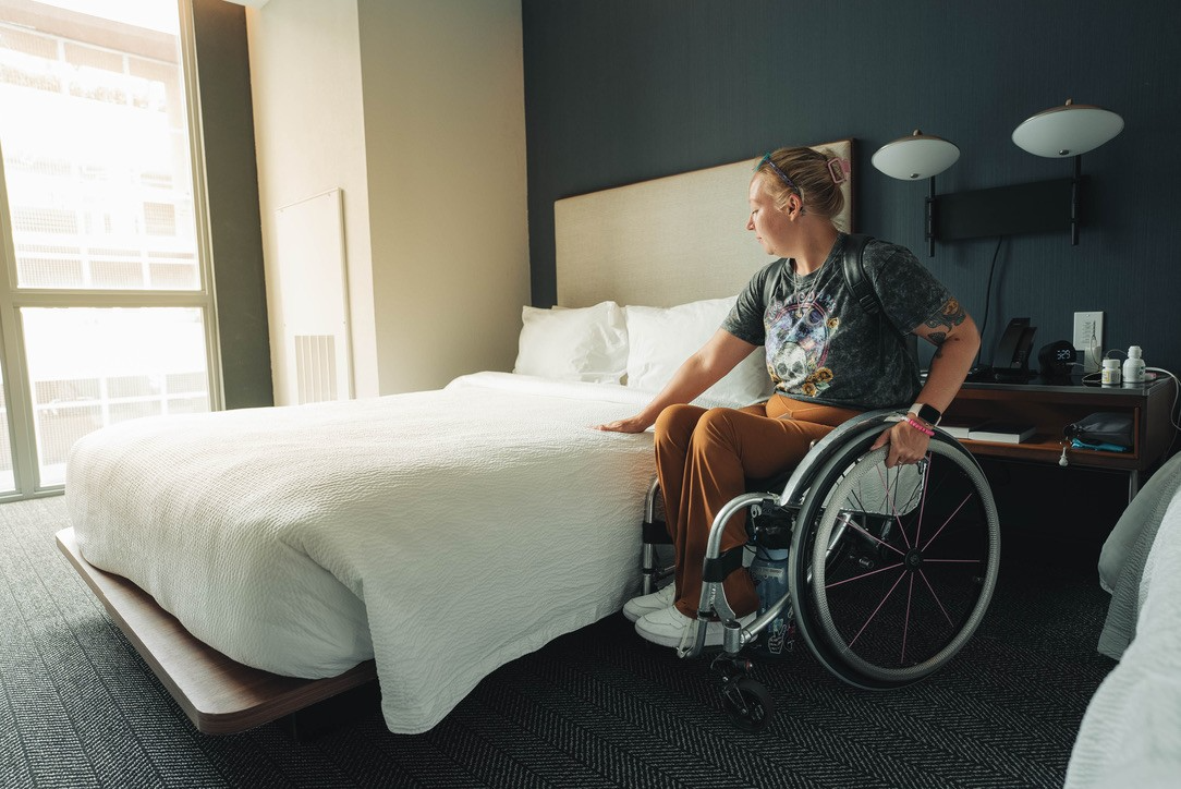 Photo of woman next to a bed in a hotel, about to transfer from her wheelchair