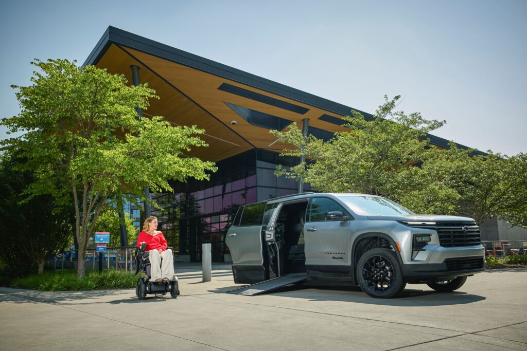 A woman in a wheelchair poses next to a Next Gen Traverse in front of a large building