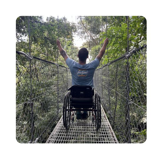 A man in a wheelchair stretching out his arms on a rope bridge surrounded by forest. For the travel hub